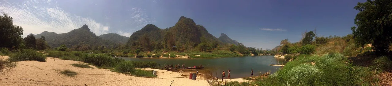 Sandbar landing on a green river beneath karst peaks, with a long-tail boat and people standing in shallow water.